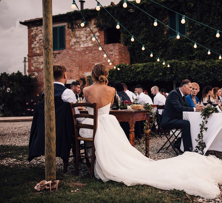 Bride in Structured Bustier Rue de Seine Fox Gown with Floral Appliqué Detail and Off The Shoulder Straps | Pronovias Chiffon Overskirt | Groom in Three-Piece Hawes &amp; Curtis Suit with Ivory Tie and Pocket Square | Festoon Lights | Outdoor Wedding Reception at Il Rigo Agriturismo, Tuscany | Rue de Seine Wedding Dress with Pronovias Overskirt, Personalised Place Setting Illustrations and Sweetheart Table | Samuel Docker Photography