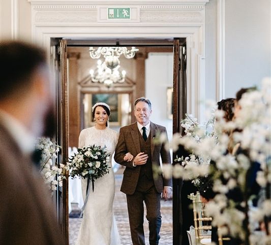 Bride walking down the aisle with her father wearing tweed wedding suit