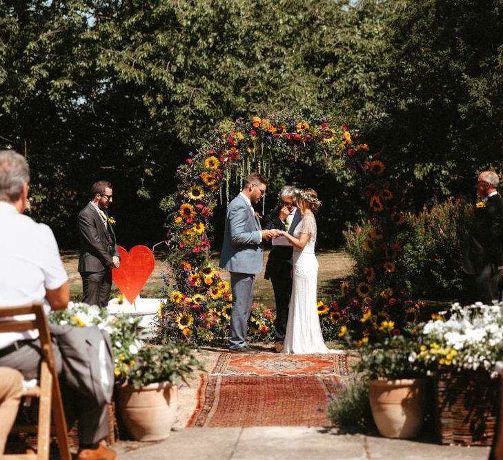 Bride in Jenny Packham Portia Wedding Dress and Groom in Grey Ted Baker Suit Exchanging Vows at Outdoor Wedding Ceremony with Floral Moon Gate,