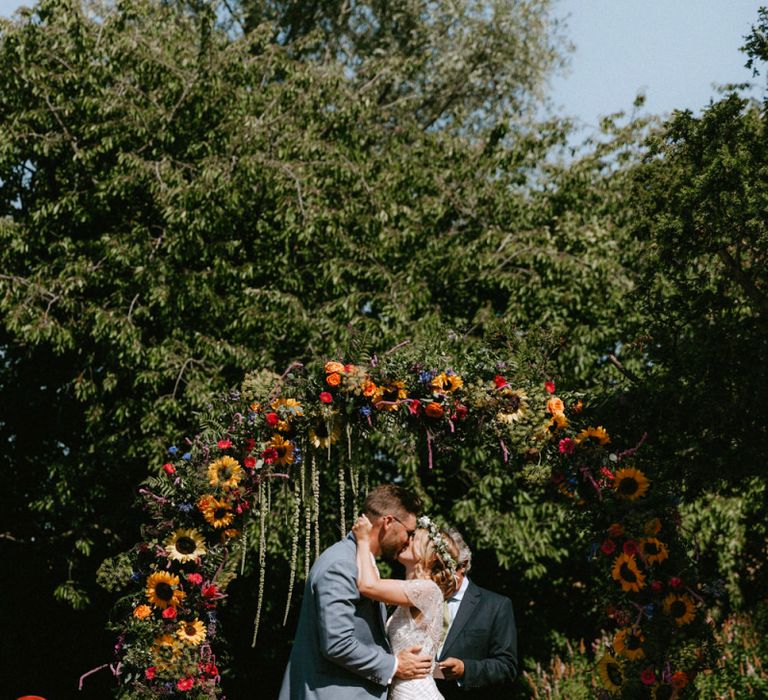 Bride in Jenny Packham Portia Wedding Dress and Groom in Grey Ted Baker Suit Kissing at the Floral Moon Gate Altar