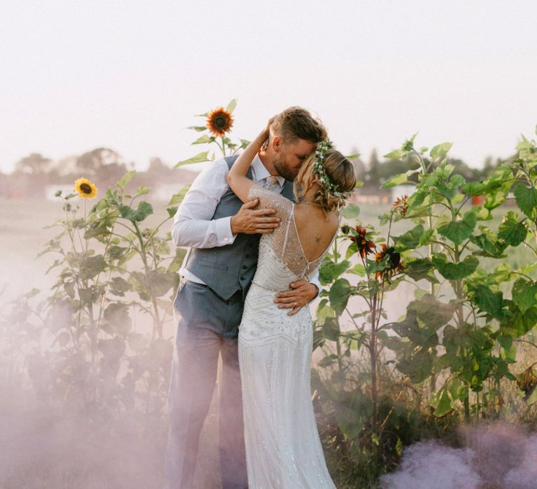 Bride in Jenny Packham Portia Wedding Dress and Flower Crown and Groom in Ted Baker Suit Kissing in a Sunflower Field with Smoke Bombs Going Off