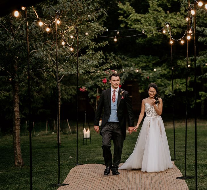 Bride and groom walking along a festoon light path to the tipi wedding reception at Evenley Wood Garden