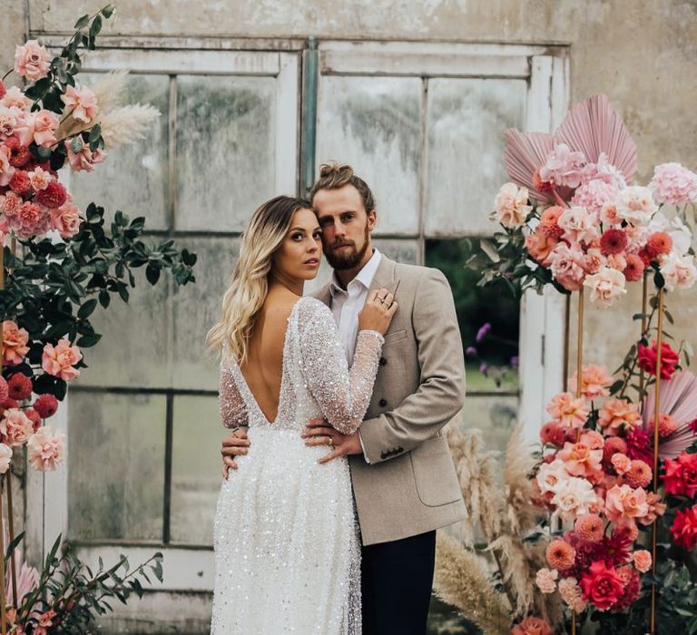 Bride in sparkle dress and groom in beige blazer surrounded by pink wedding flowers