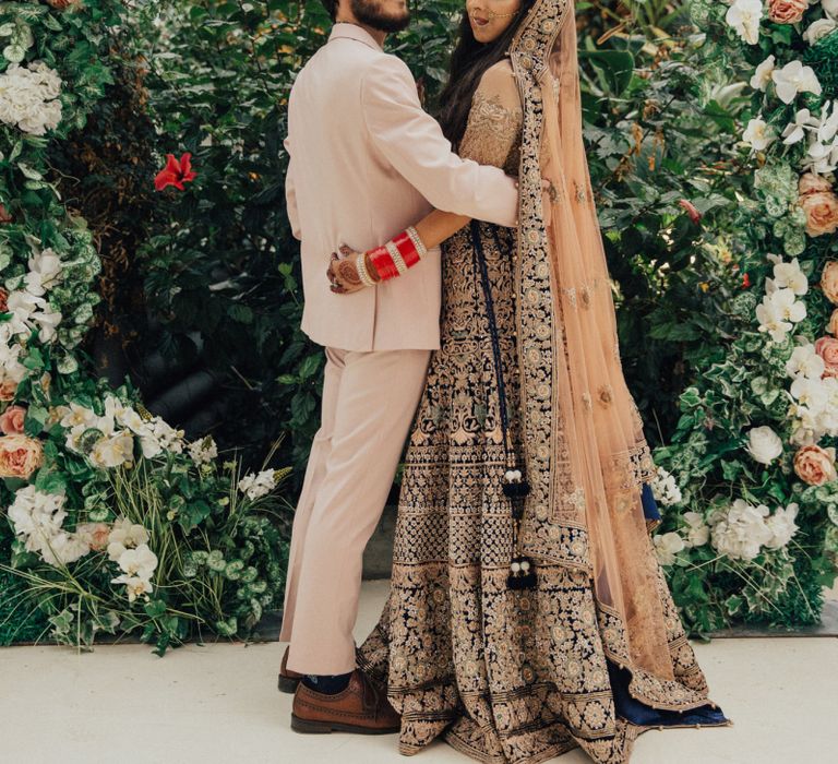 Bride in Gold Saree and Groom in Beige Suit Standing in Front of Floral Moon Gate