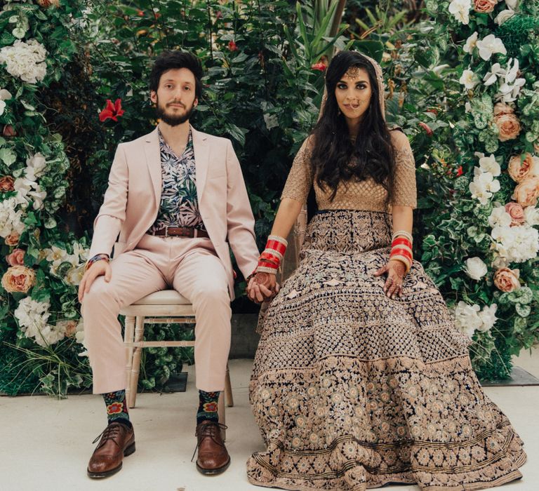 Bride in Gold Saree and Groom in Beige Suit Sitting in Front of Floral Moon Gate