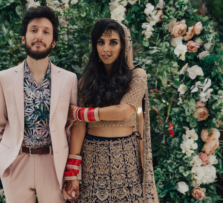 Bride in Gold and Navy Saree and Groom in Beige Suit and Floral Shirt Arm in Arm in Front of Floral moon Gate