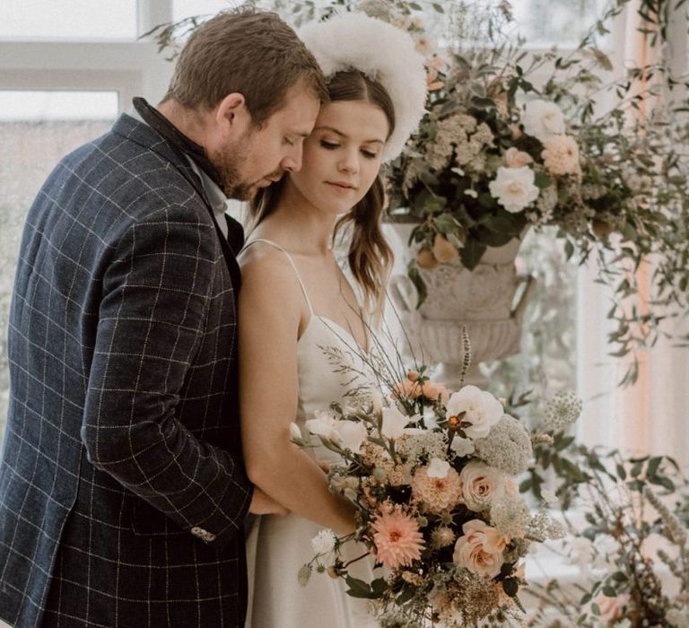 Groom in Blue Checked Blazer Embracing His Bride in an Emma Beaumont Silk Wedding Dress at the Floral Altar