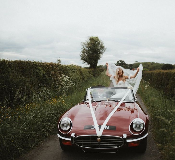 Bride in Emma Beaumont Wedding Dress Sits on Convertible Wedding Car While Groom Drives Down a Country Lane