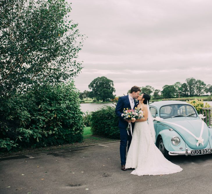 Bride in Off The Shoulder Wedding Dress and Groom in Navy Suit Kissing By Blue VW Beetle Wedding Car