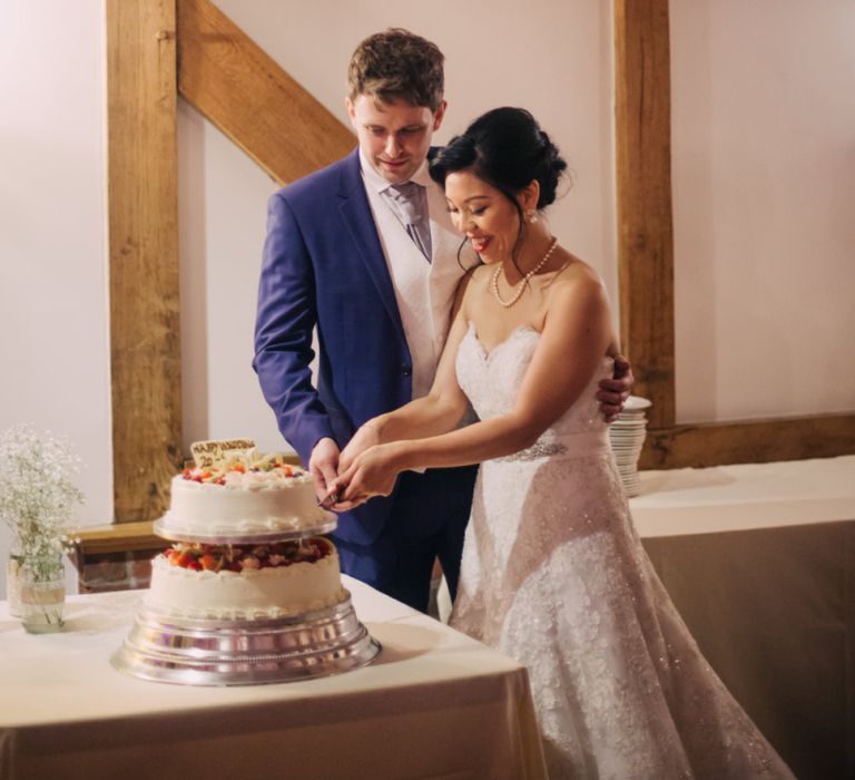 Bride in Lace Off The Shoulder Wedding Dress and Groom in Tuxedo Cutting The Wedding Cake