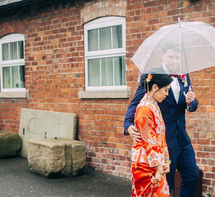 Groom in Black Tuxedo Holding umbrella over bride in red Chinese wedding dress