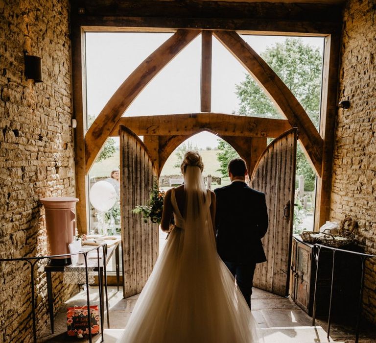 Bride in Stella York Wedding Dress and Cathedral Length Veil and Groom in Navy Suit Standing at the Entrance to Cripps Barn