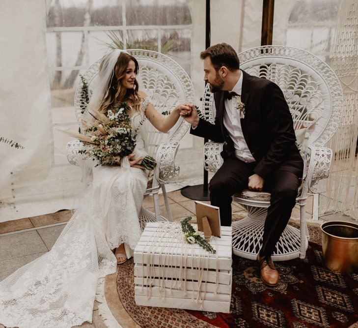 Bride and groom in brown wedding suit sit on peacock chairs during ceremony