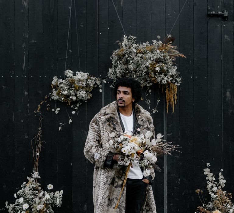 Stylish Groom in White T-Shirt and Faux Fur Overcoat Holding a Dried Flower Bouquet