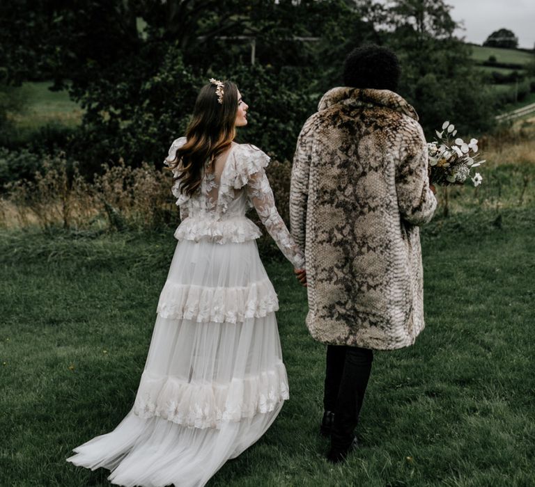 Bride in Tiered Wedding Dress and Groom in Faux Fur Coat Walking Through the Countryside