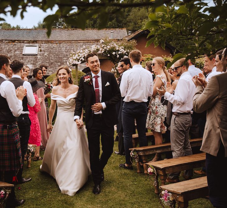 Bride in Bardot Art Couture Wedding Dress and Groom in Black Moss Bros. Suit with Burgundy Tie Walking Up the Aisle as Husband and Wife