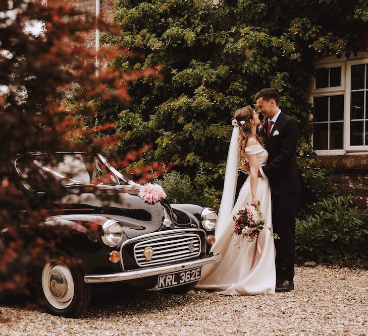 Bride in Bardot Wedding Dress and Groom in Black Moss Bros. Suit and Burgundy Tie Standing Next to their Vintage Black Wedding Car