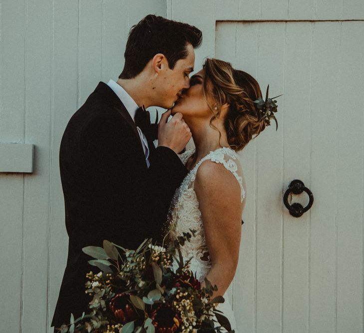 Bride in The Eleni Wed2B Lace Wedding Dress Holding a Protea Bridal Bouquet and Groom in Tuxedo Kissing