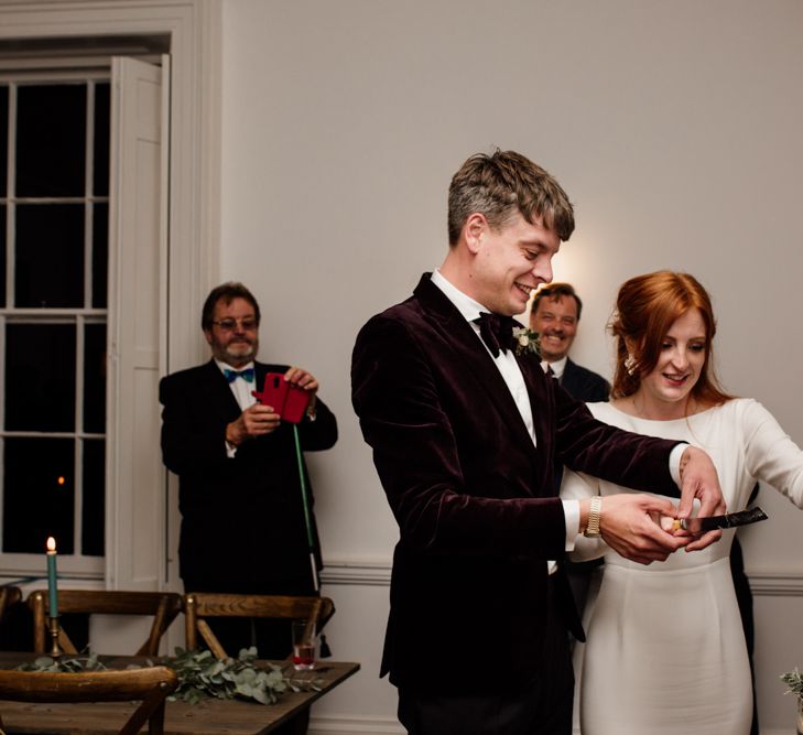 Groom in velvet tuxedo and bride in minimalist wedding dress cutting the wedding cake