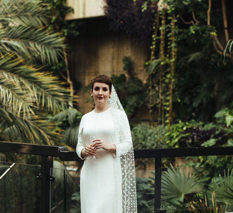 Bride  with Pixie Cut Hair in Charlie Brear Minimalist Wedding Dress with Long Sleeves and Front Split and Polka Dot Wedding Veil