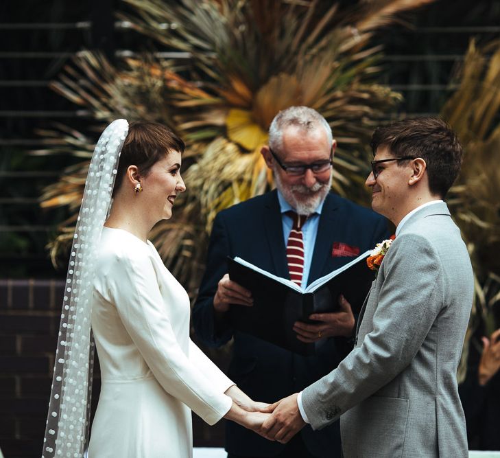 Bride in Minimalist Charlie Brear Wedding Dress with Long Sleeves and Groom in Grey Suit Holding Hands During the Wedding Ceremony