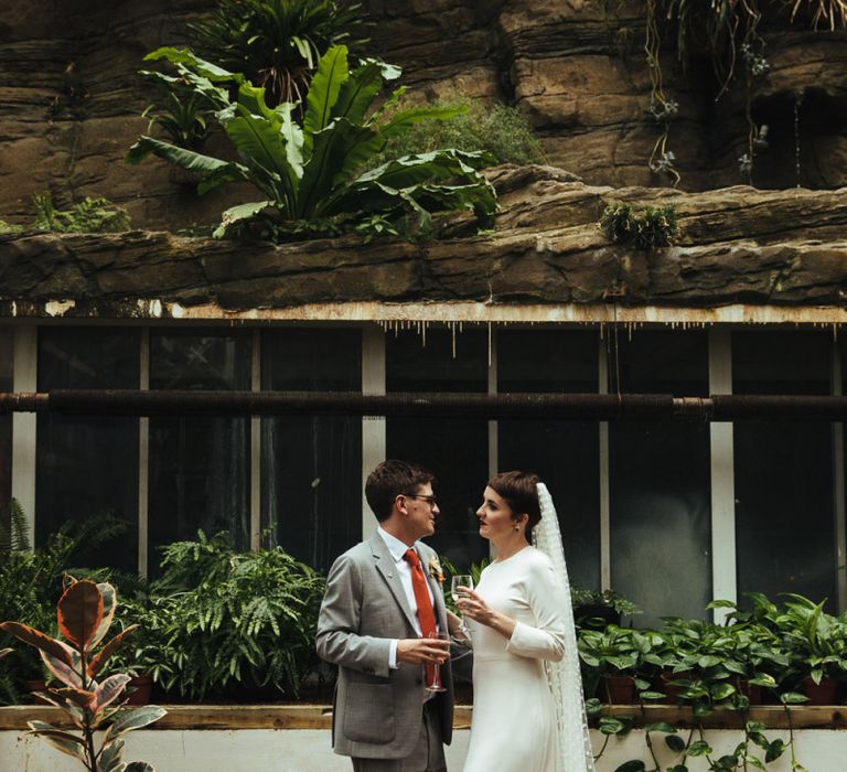 Bride in Charlie Brear Wedding Dress and Polka Dot Wedding Veil and Groom in Light Grey Suit Enjoying a Glass of Champagne Together