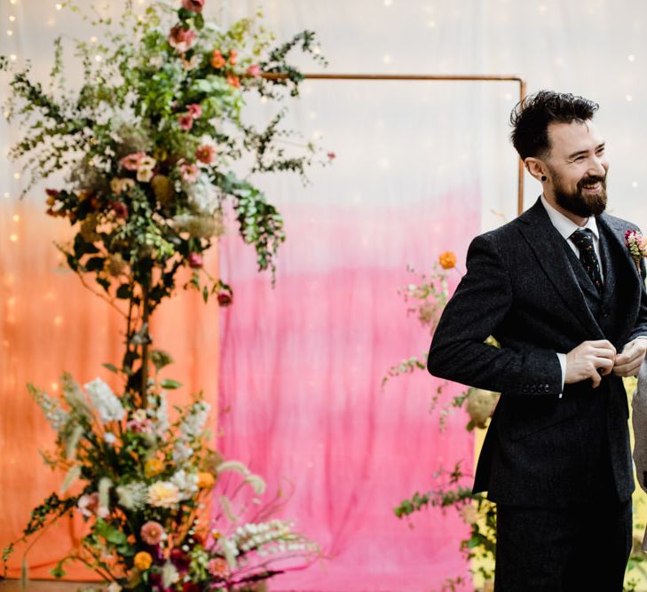 Groom at the top of the aisle with pink, orange and yellow backdrop and copper archway decorated with floral decor