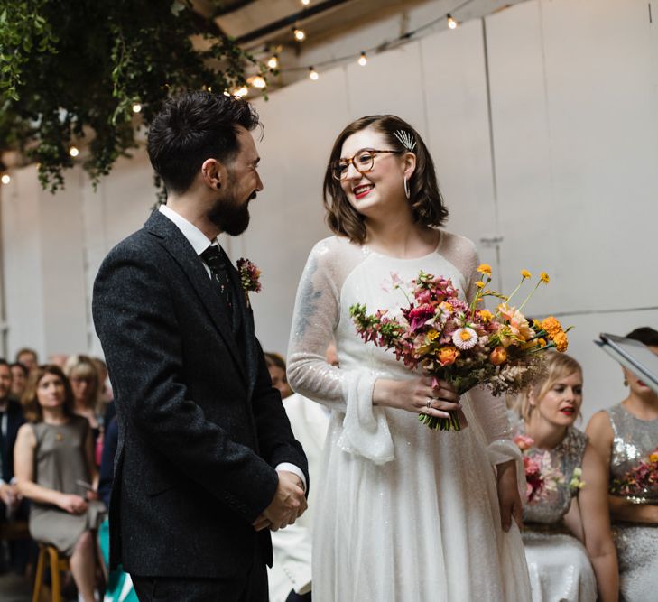 Bride in glasses at industrial wedding ceremony with bright floral bouquet and hanging festoon lighting