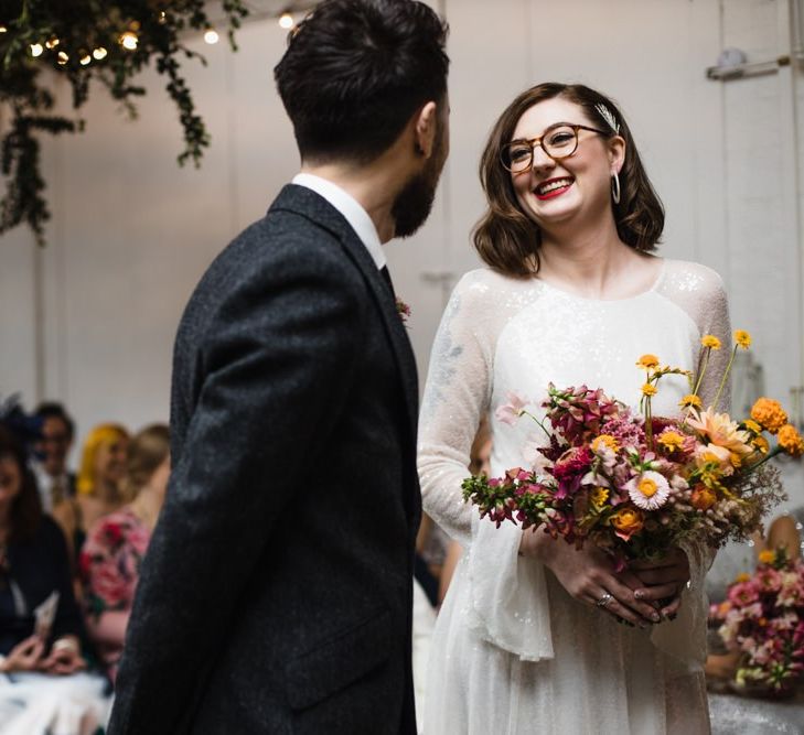 Bride in glasses at industrial wedding ceremony with hanging festoon lighting and bright floral decor