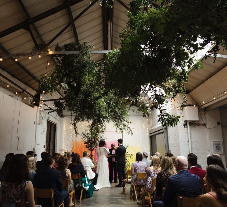 Bride in glasses and groom tie the knot at industrial wedding ceremony with hanging foliage and festoon lighting and