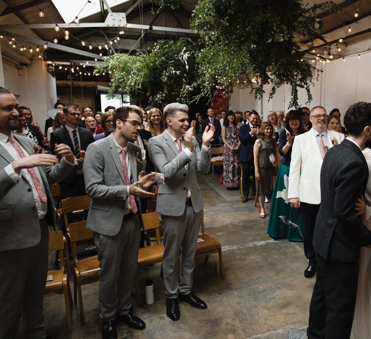 Bride in glasses and groom say 'I do' with their guests at industrial wedding ceremony with festoon lighting and hanging foliage decor