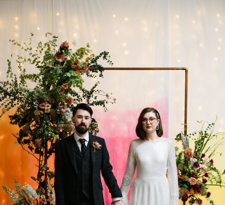 Bride in glasses and groom stood by bright hanging backdrop with copper archway and floral decor