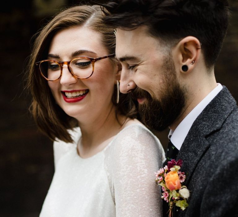 Bride in glasses and long embellished sleeve dress with groom wearing purple and orange floral buttonhole