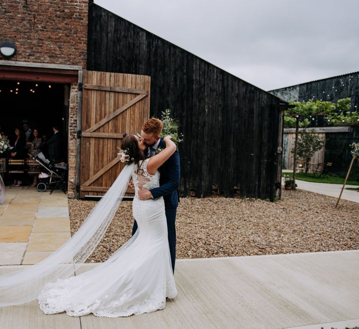 Bride in Lace Roland Joyce Bridal Wedding Dress and Groom in Navy Suit Kissing Outside Barn Wedding Venue