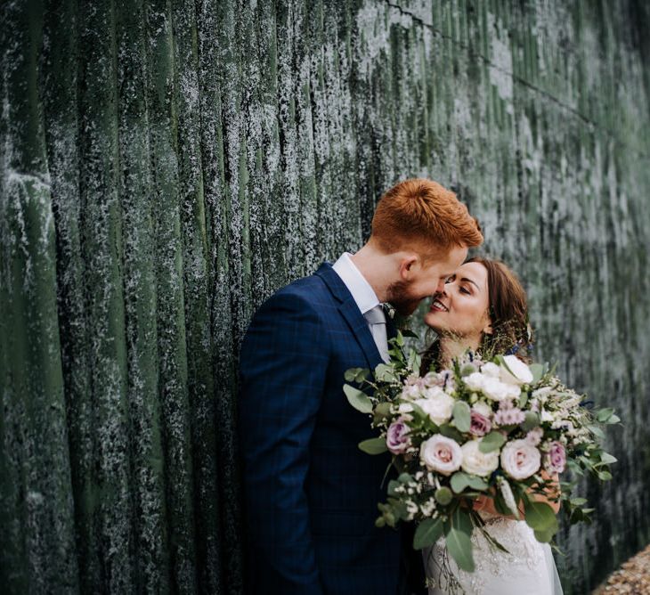 Bride in Lace Roland Joyce Bridal Wedding Dress Holding a Pink Rose Bouquet Kissing Her  Groom in a Navy Suit