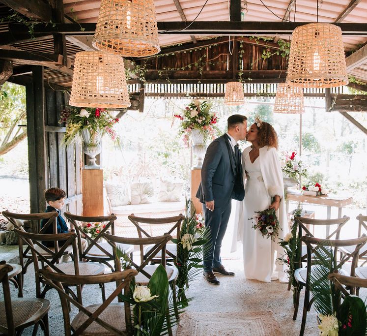 Bride with Afro Hair in Gold Headdress, Patricia Meléndez Wedding Dress and coat and Groom in Navy Suit Kissing in wooden ‘pagoda’ decorated with wicker lamps and botanical plants