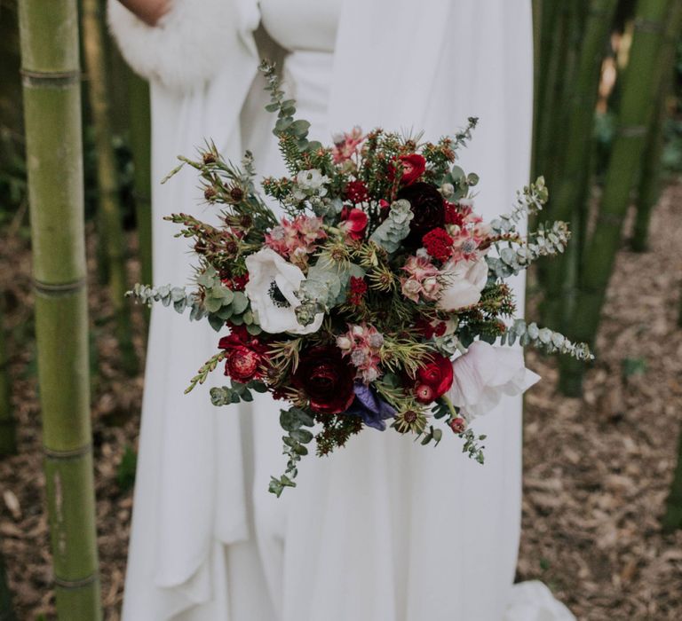 Bride in Patricia Meléndez Wedding Dress and Coat with blue fox wristband Holding Green, Red and White Bouquet