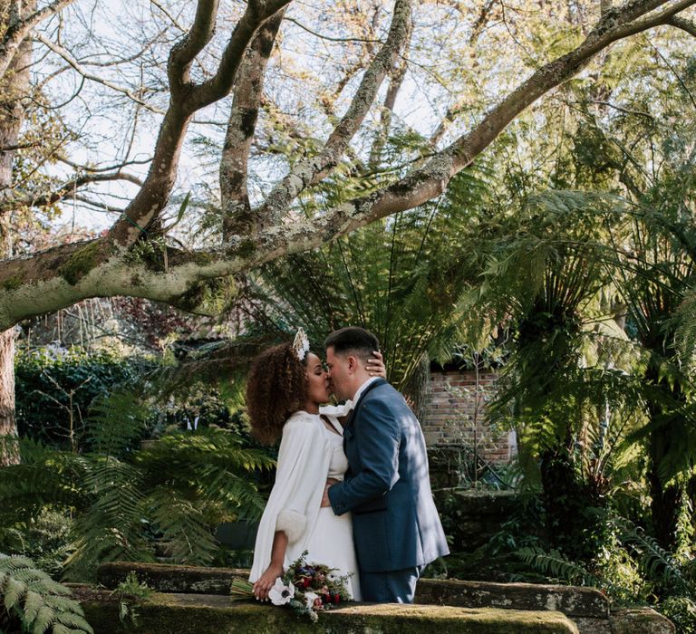 Bride in Patricia Meléndez Wedding Dress and Coat with blue fox wristband and Groom in Navy Suit Kissing