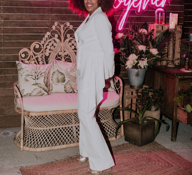 Bride in White Trouser Suit Standing in Front of Chill-out Zone with Wicker Chair and Pink Neon Better Together Sign