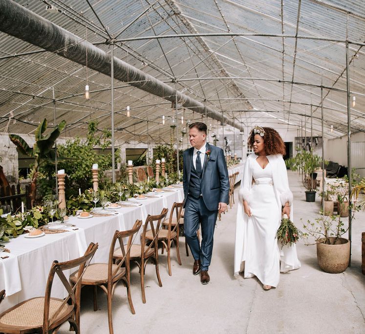 Bride with Afro Hair in Gold Headdress, Patricia Meléndez Wedding Dress and coat and Groom in Navy Suit Walking Through Glasshouse Wedding Venue