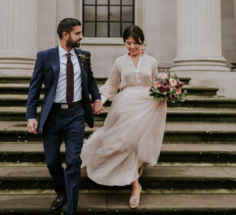 Bride and groom walking down the steps at The Old Marylebone Town Hall in a tulle skirt and navy suit