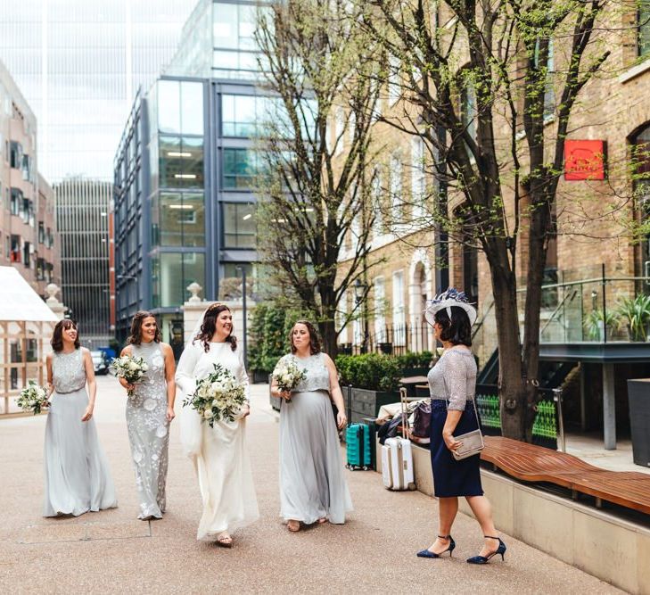 Bride arriving with her bridesmaids to courtyard ceremony wearing soft grey dresses with white floral bouquets