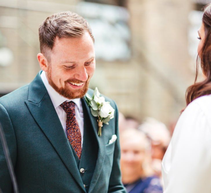 Groom wearing green suit with white floral buttonhole at courtyard ceremony with foliage decor