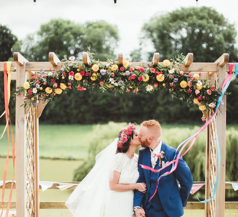 Flower Covered Arbour | Bride in Lace Watters Gown &amp; Colourful Flower Crown | Groom in Navy Suit | Bright Festival Themed Outdoor Ceremony &amp; Tipi Weeding |  Maryanne Weddings | Framed Beauty Film