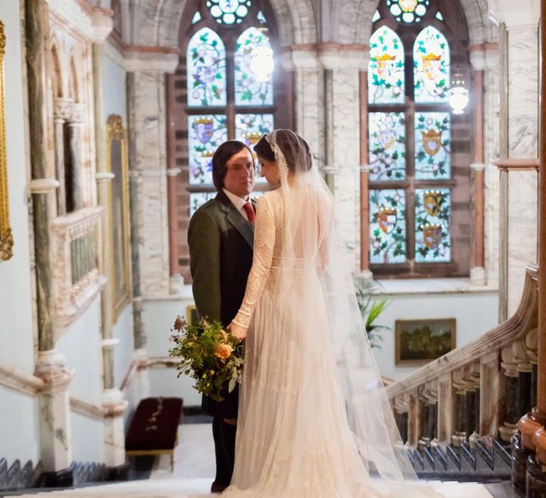 Bride in Delicate Lace Lihi Hod Sophia Wedding Dress with Long Sleeves and Groom in Traditional Tartan Kilt Standing in the Grand Hall of Mount Stuart in Scotland