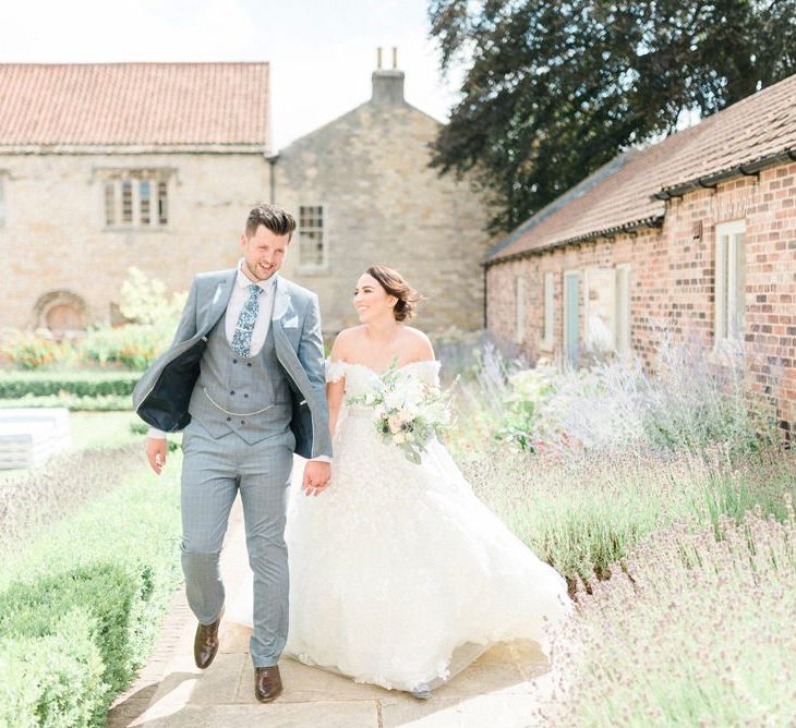 Bride in Appliqué Ian Stuart Wedding Dress and Groom in Most Suitable Blue Check Wedding Suit Walking Through The Gardens