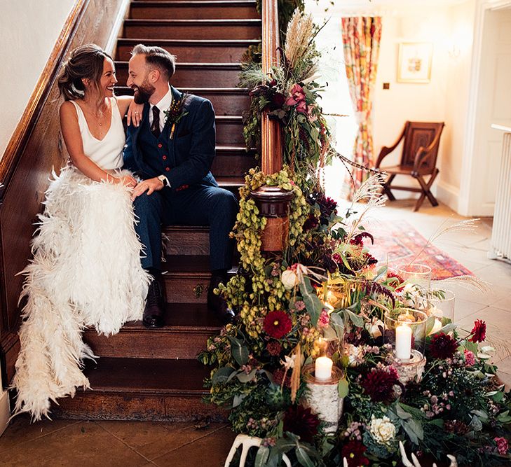 Bride Sitting on The Stairs  in a Charlie Brear Payton Bridal Gown &amp; Piora Feather Skirt with Her Groom in a Navy Suit