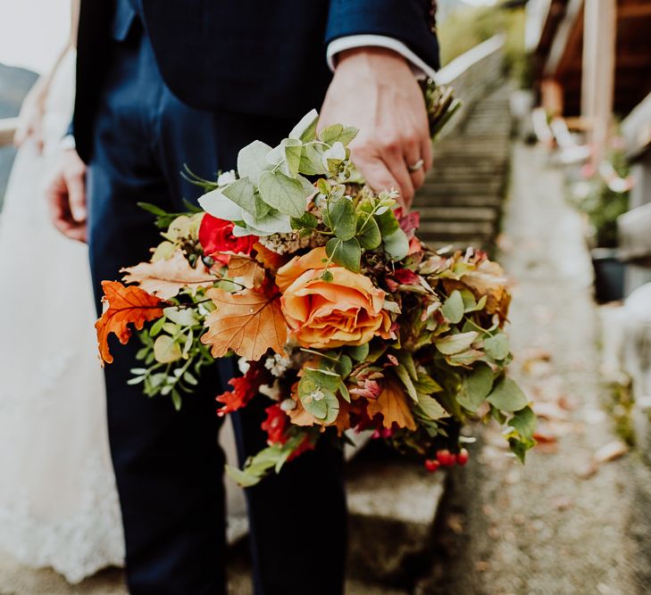 Groom Golding the Autumnal Orange Flower and Green Leaf Wedding Bouquet