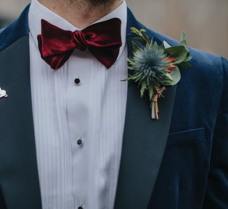 Navy velvet grooms suit details with red bow tie and festive buttonhole foliage for winter wedding