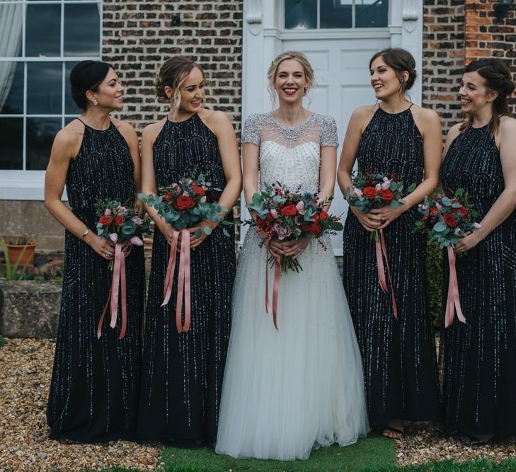 Bride wearing a beautiful embellished wedding dress and a classic red lip with her bridesmaids in black dresses and festive bouquets